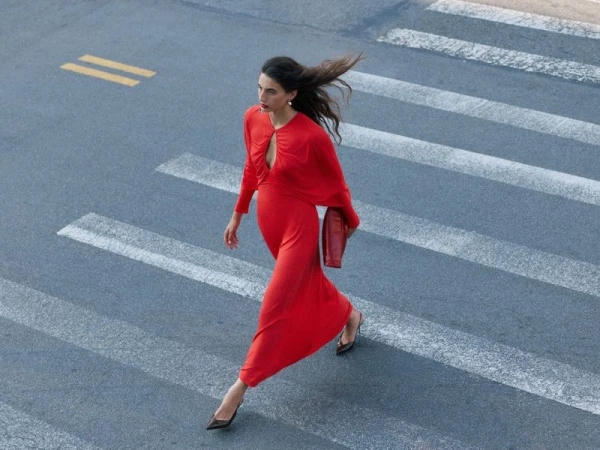 Model wearing a vibrant red dress walking across an asphalt crosswalk holding a red clutch