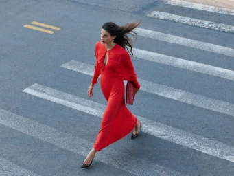 Model wearing a vibrant red dress walking across an asphalt crosswalk holding a red clutch