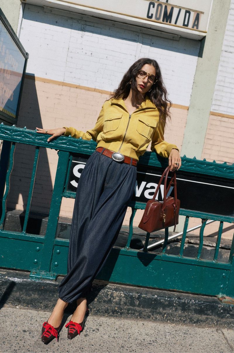 Model leaning against a green subway railing wearing a yellow jacket, dark trousers, and holding a brown handbag