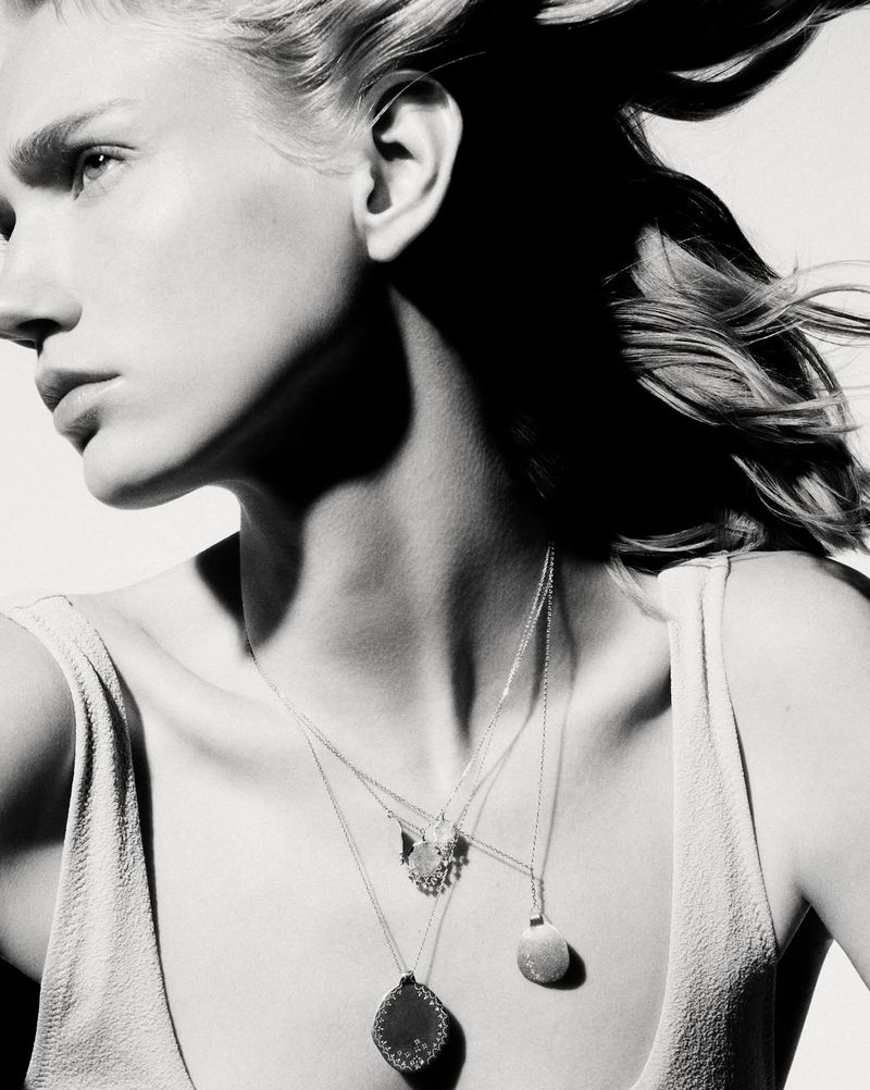 Close-up portrait of a model wearing layered pendant necklaces and a light-colored top against a bright white background.