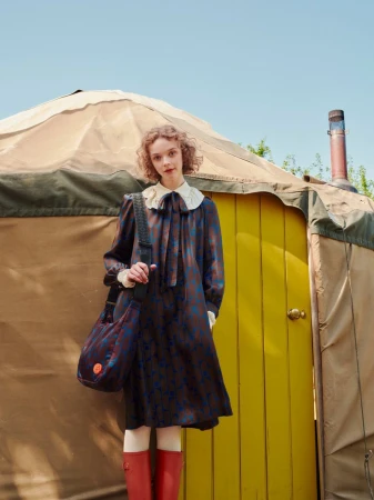 Woman in patterned dress with white collar and red boots holding a shoulder bag in front of a tent
