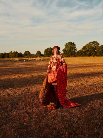 Model standing in a dry grassy meadow holding a red quilted blanket and matching patterned bag for Orla Kiely AW26.