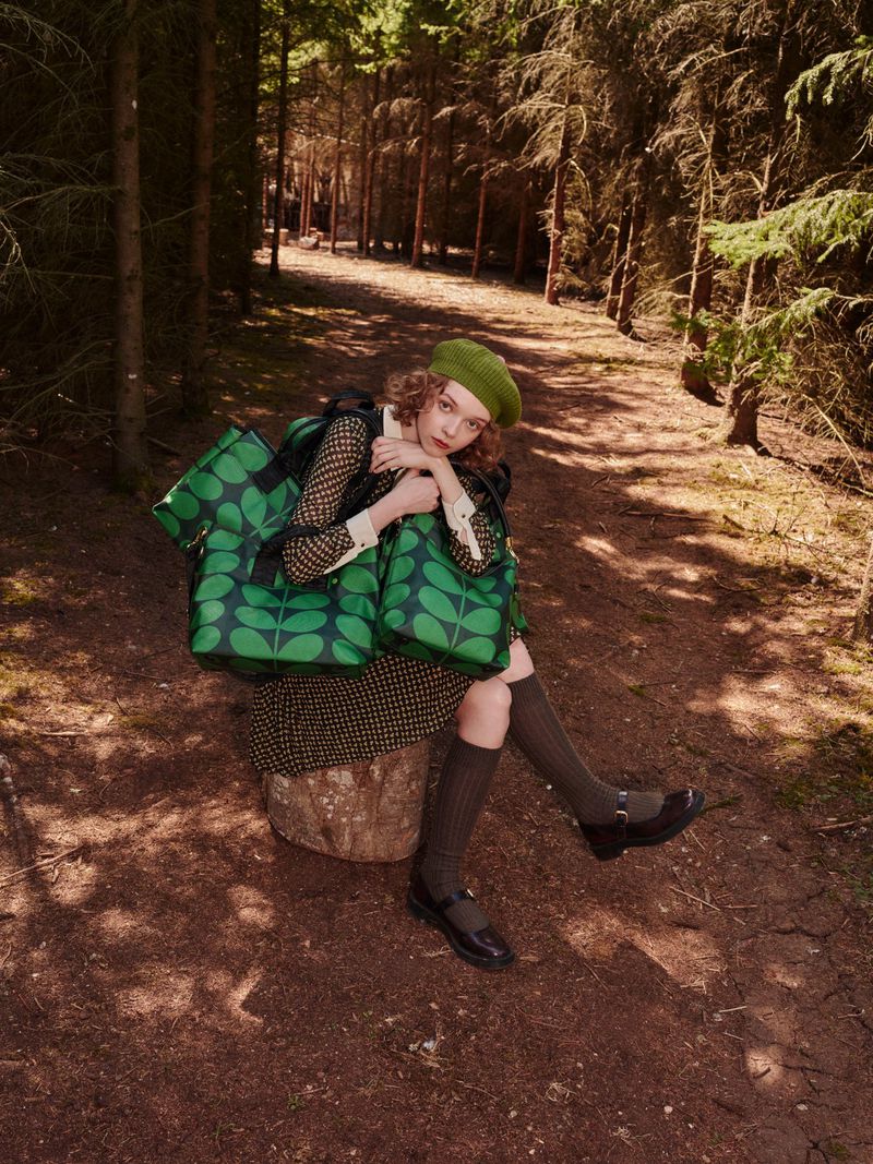 A woman wearing a green beret and patterned dress sits on a tree stump in a forest, holding several green patterned tote bags.
