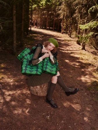 A woman wearing a green beret and patterned dress sits on a tree stump in a forest, holding several green patterned tote bags.
