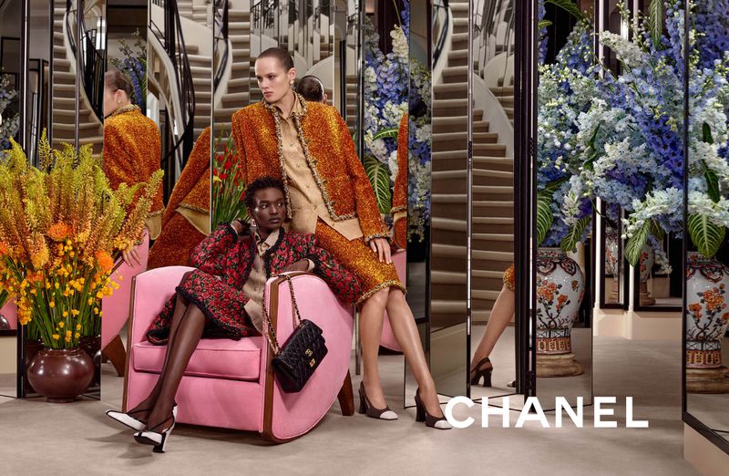 Two models wearing Chanel tweed suits in a mirrored salon with floral arrangements and a pink armchair