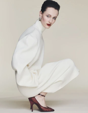 Model crouching against a neutral background wearing a cream turtleneck dress and dark burgundy heels.