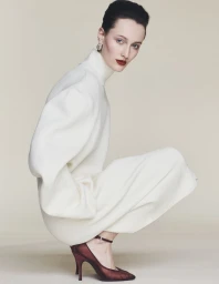 Model crouching against a neutral background wearing a cream turtleneck dress and dark burgundy heels.