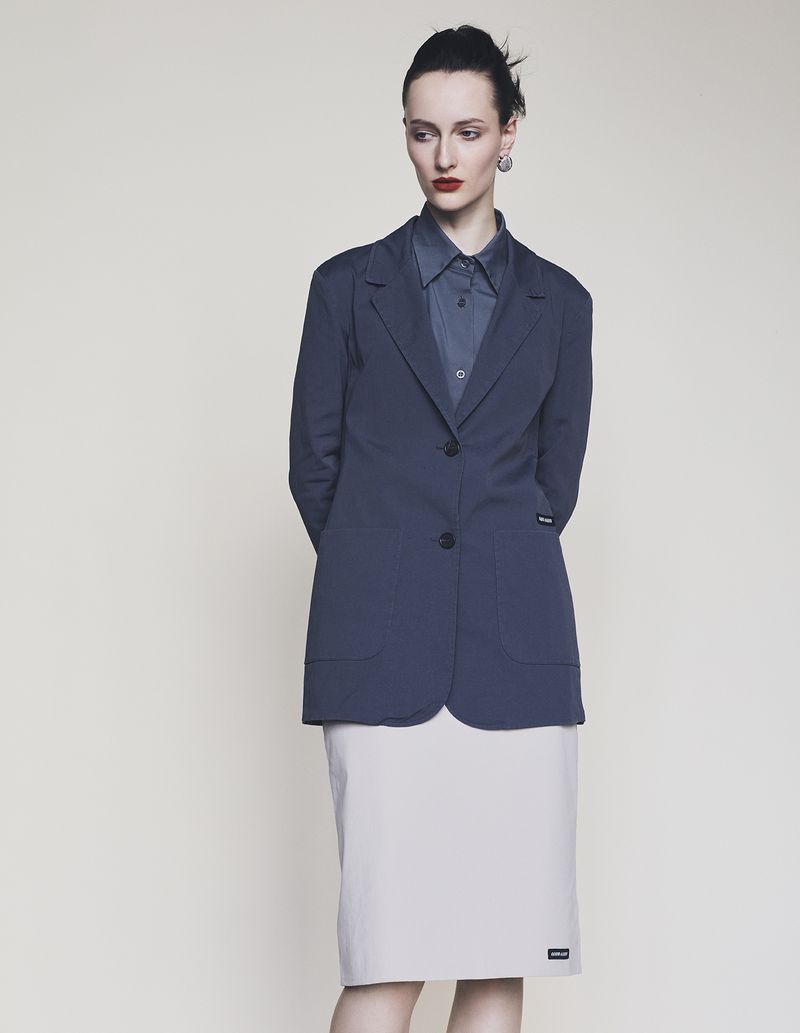 Model wearing a dark blazer and light skirt standing against a neutral background for Harper's Bazaar Germany.