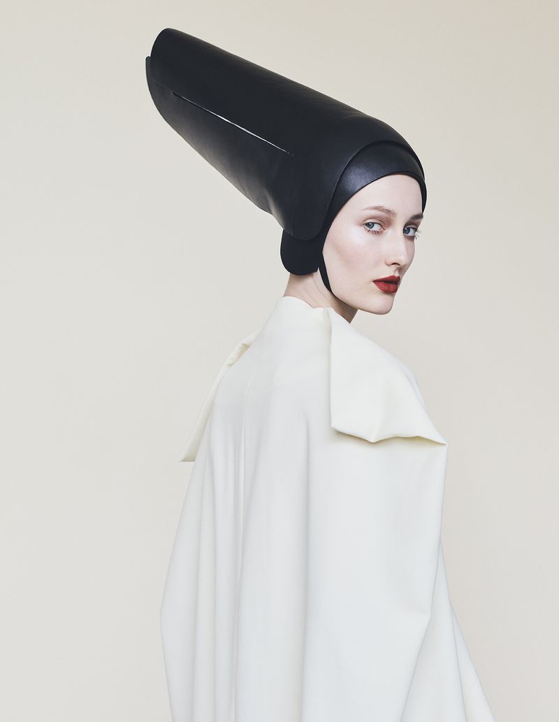 Model wearing a tall structured black headpiece and white high-collared coat against a seamless light background for Harper's Bazaar Germany.