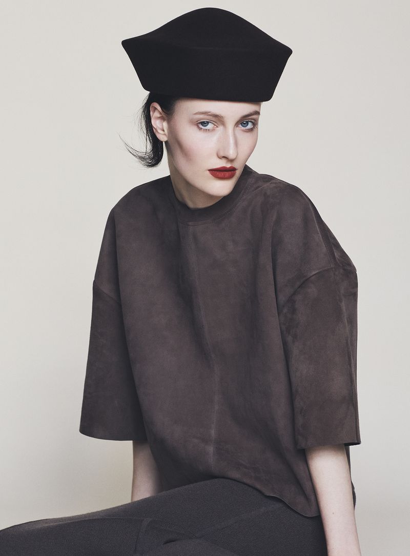 Model wearing a structured black cap and dark brown top against a neutral off-white background