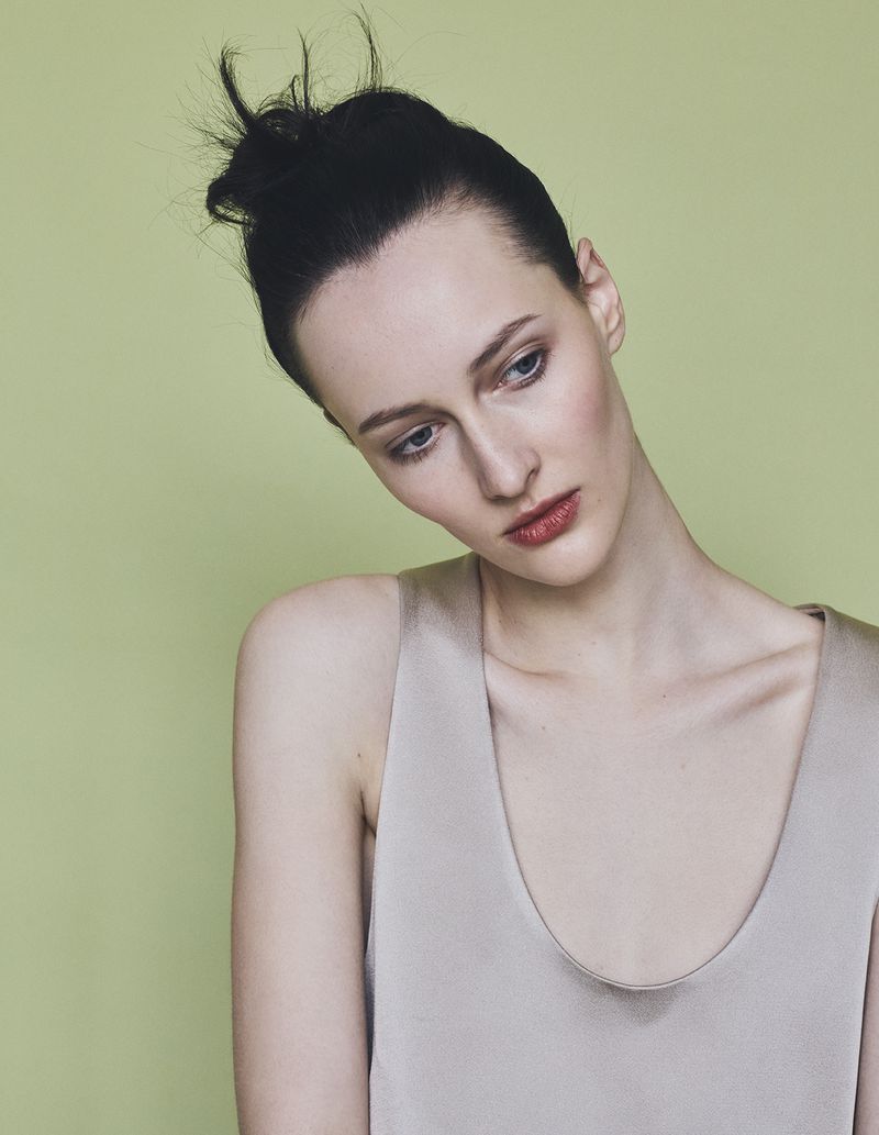 Model with dark hair in a high bun wearing a grey top against a muted green studio backdrop for Harper's Bazaar Germany