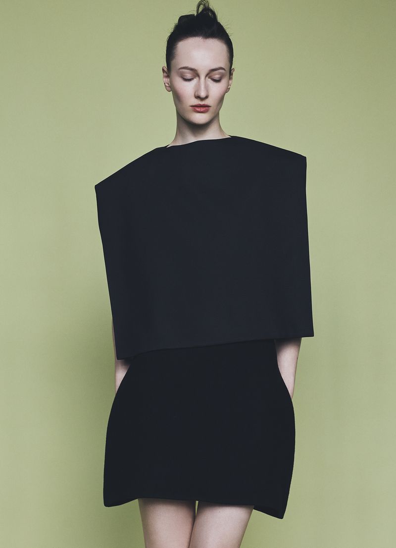 Model wearing a structured black architectural top and skirt against a solid green background