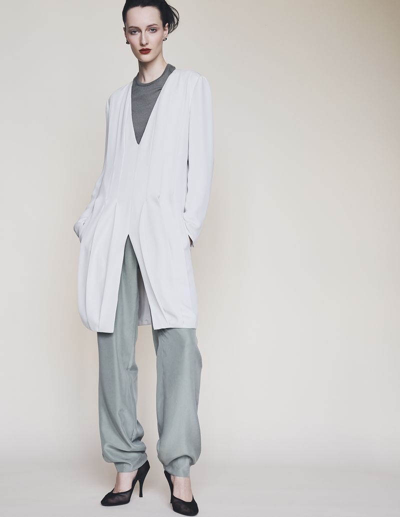 Model Mauve Dupuy wearing a long white coat over grey trousers against a plain studio background.