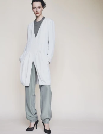 Model Mauve Dupuy wearing a long white coat over grey trousers against a plain studio background.