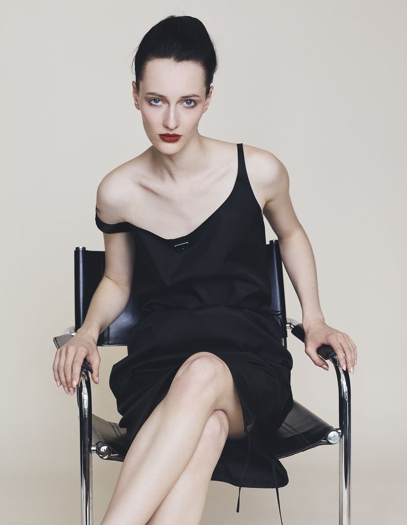 Model wearing a black slip dress seated in a leather chair against a white background