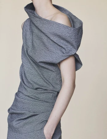 Model wearing a grey textured sculptural asymmetrical draped top against a plain beige background