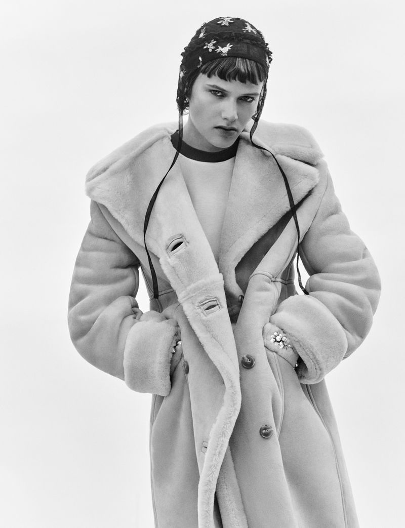 Model wearing a thick shearling coat and embroidered cap against a neutral background in a black and white fashion photograph.