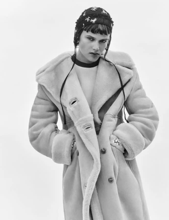 Model wearing a thick shearling coat and embroidered cap against a neutral background in a black and white fashion photograph.