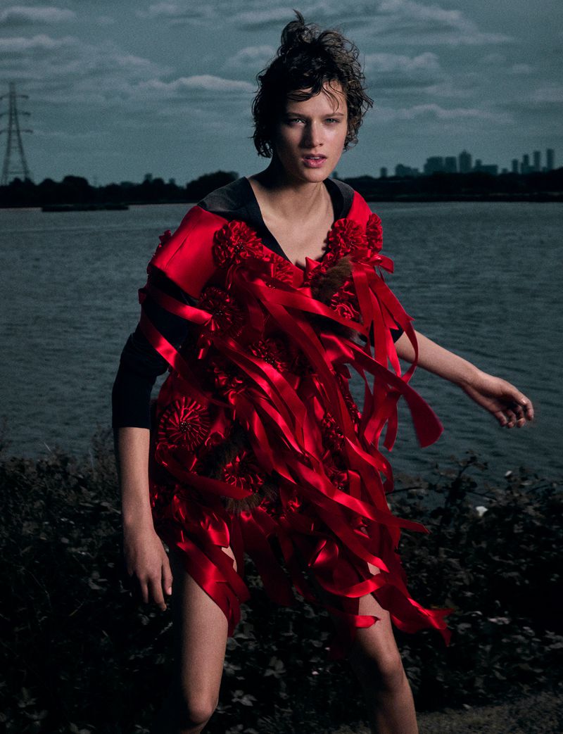 Model in a red ribbon-adorned dress standing by a waterfront with a distant cityscape under an overcast sky