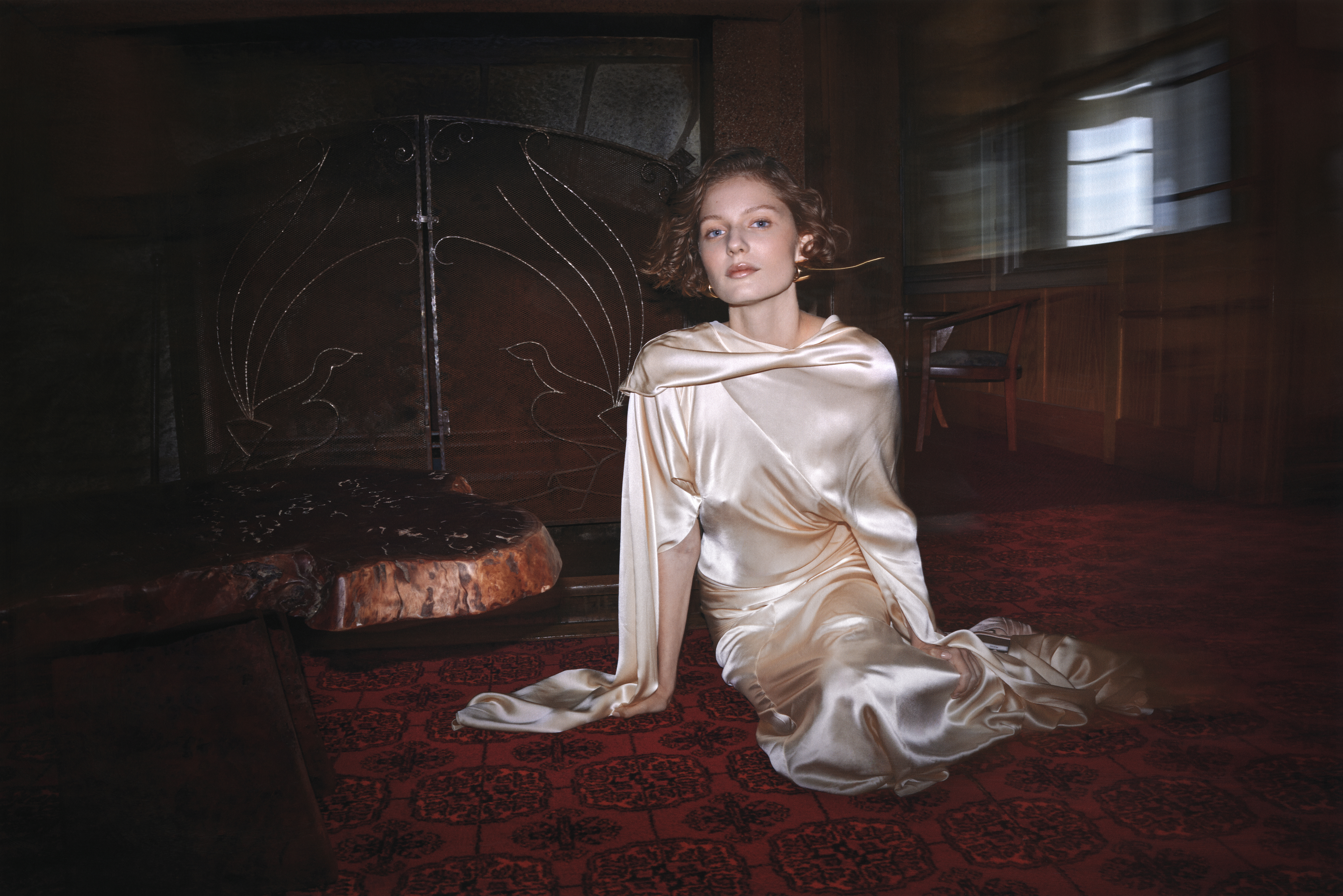 Adult female in draped satin gown sits on patterned red carpet in a blurred, warmly lit room with an ornate metal fireplace screen.