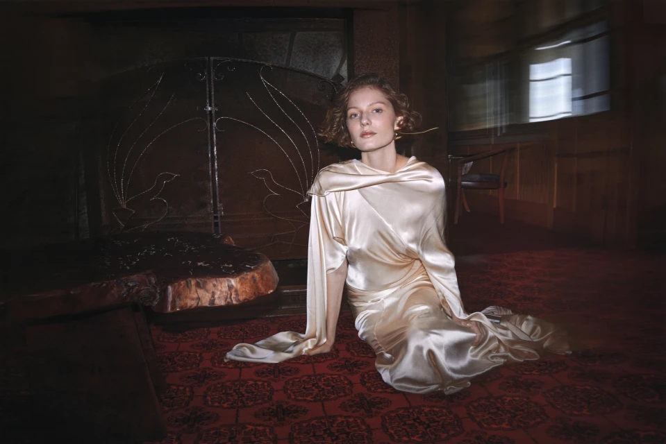 Adult female in draped satin gown sits on patterned red carpet in a blurred, warmly lit room with an ornate metal fireplace screen.