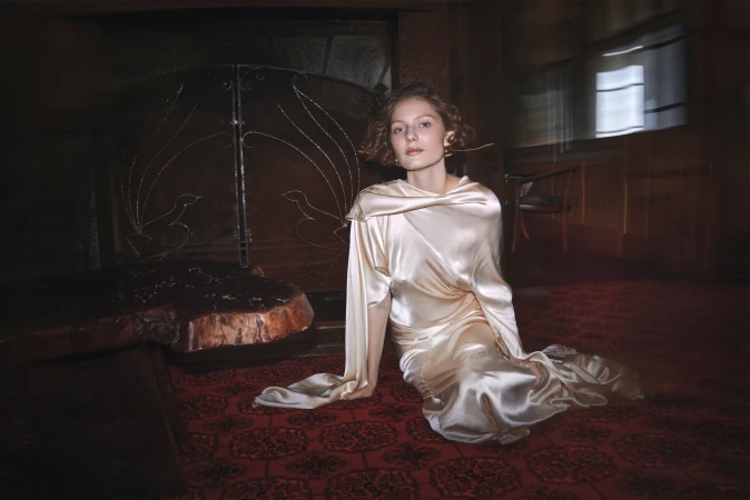 Adult female in draped satin gown sits on patterned red carpet in a blurred, warmly lit room with an ornate metal fireplace screen.