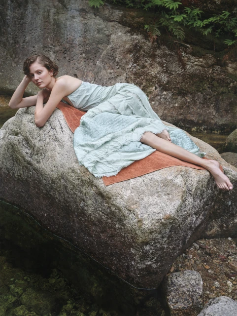 Woman in light blue dress lying on a large rock in a riverbed, surrounded by natural greenery and textured rocks, fashion editorial.