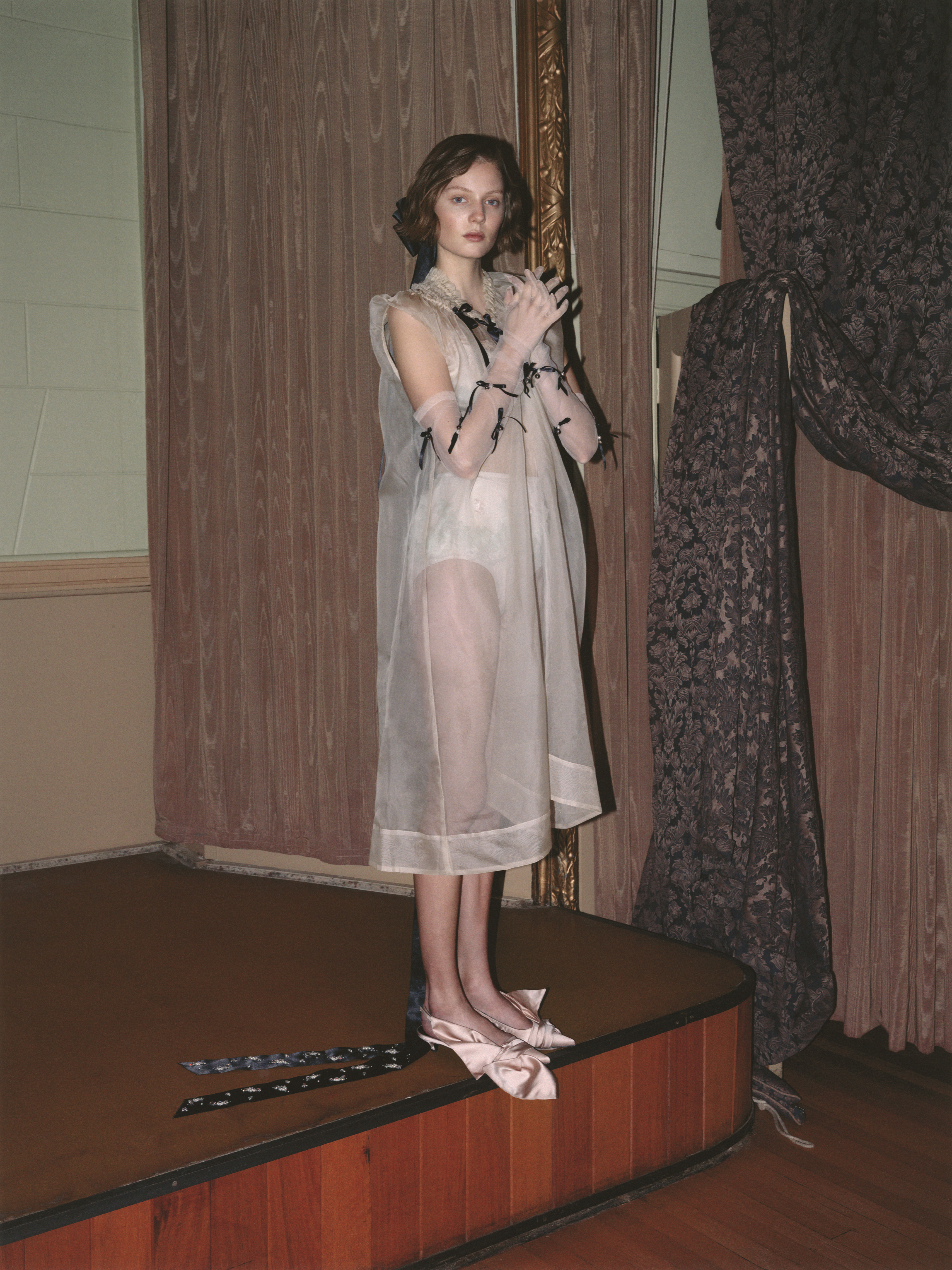 Woman stands on a stage in a sheer dress with ribbons on her arms, wearing ballet slippers with bows, with an ornate curtain behind.