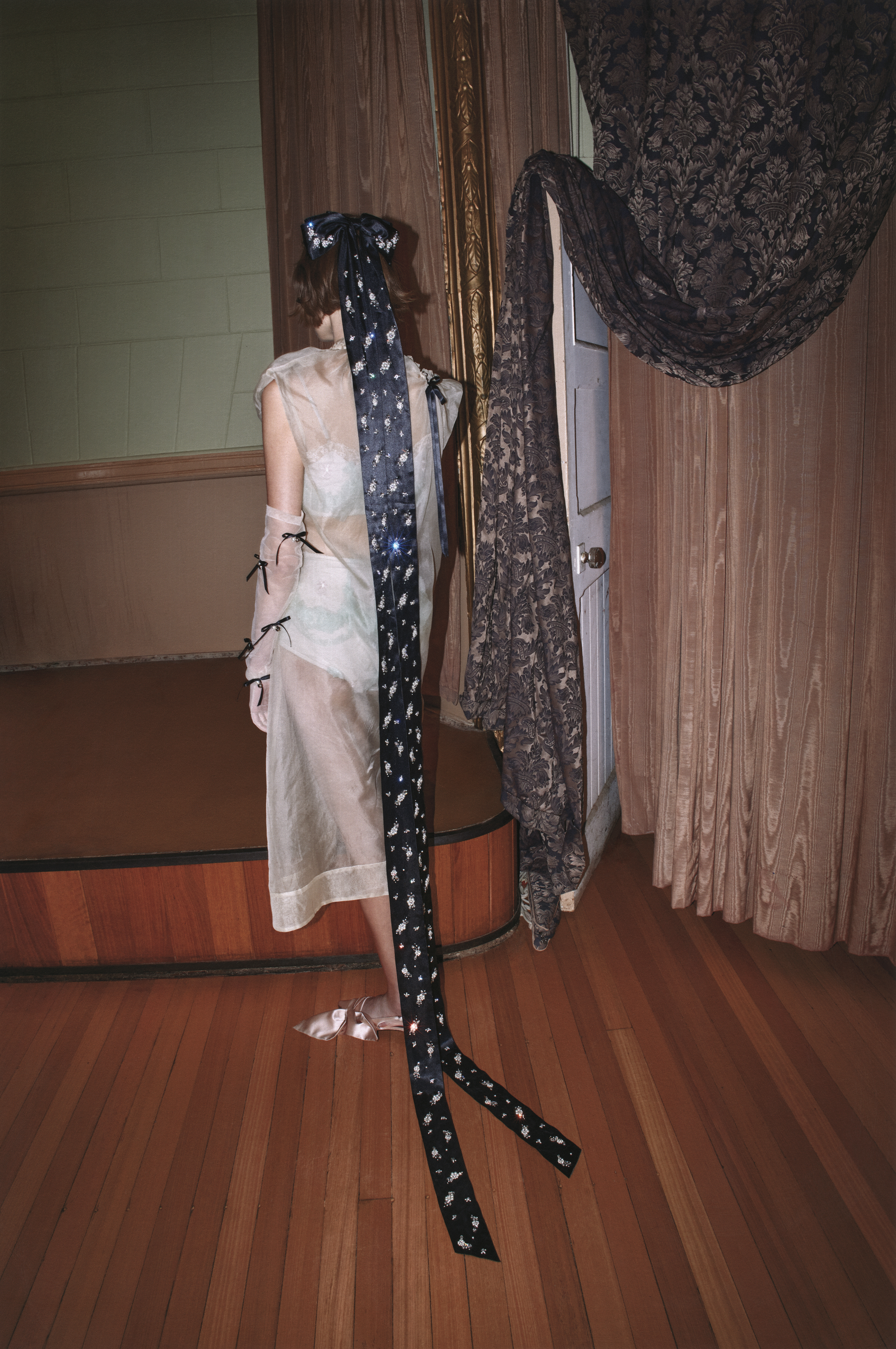 A woman in a sheer dress poses indoors with dramatic headpiece, gloves, and curtain, highlighting fashion and design elements.
