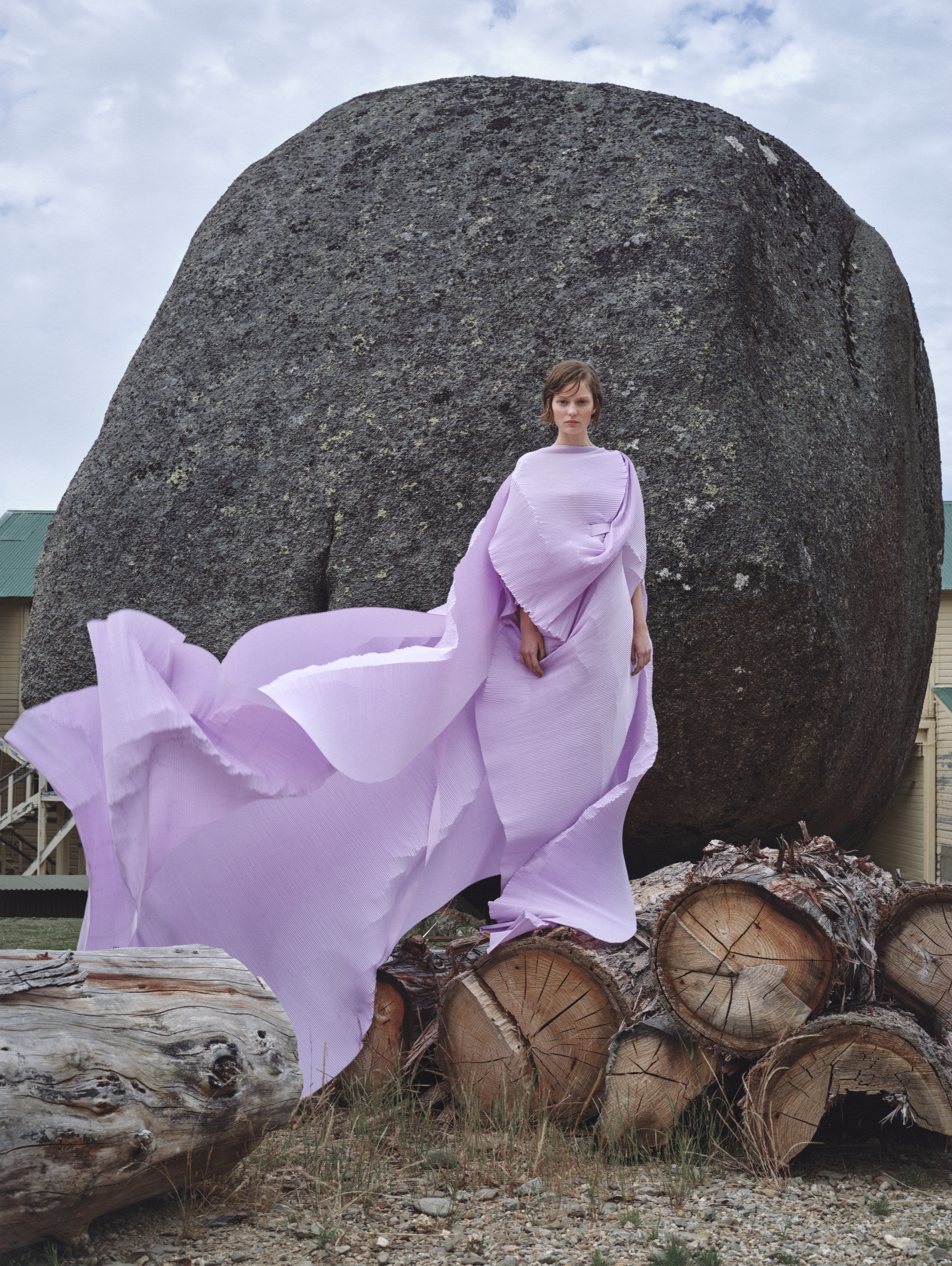 A woman in a flowing lilac gown poses near a large boulder and stacked logs in a rural setting under a cloudy sky, fashion editorial.