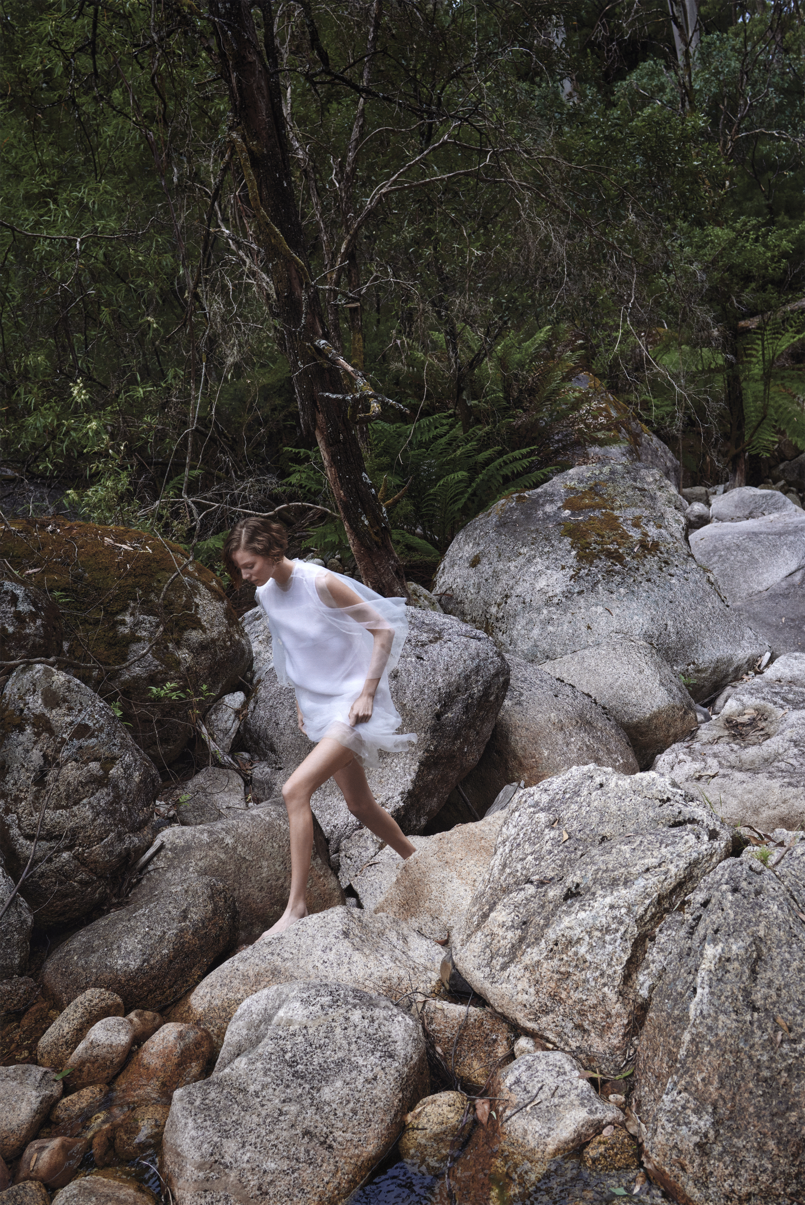 Model in sheer white dress stepping across large grey river boulders in a lush forest