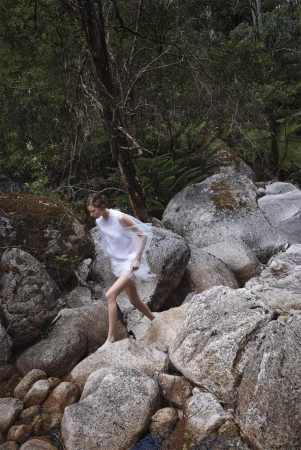 Model in sheer white dress stepping across large grey river boulders in a lush forest