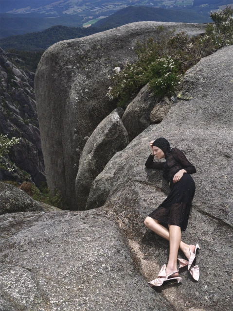 Fashion editorial shot of a woman lying on rocks with a dress and cap with mountains and trees in the background. 