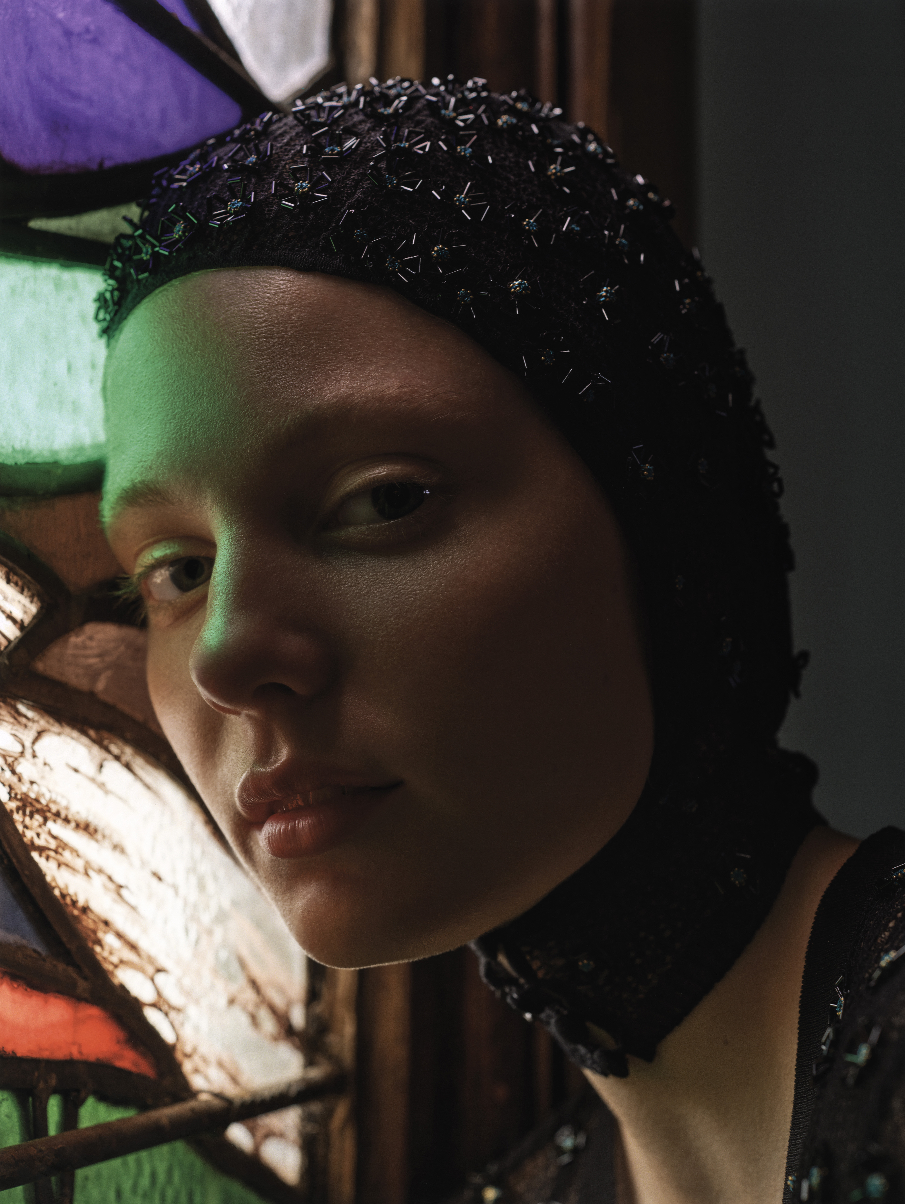 Close-up of a woman wearing a black beaded cap and choker, posing near a stained-glass window, creating a unique, intimate fashion portrait.