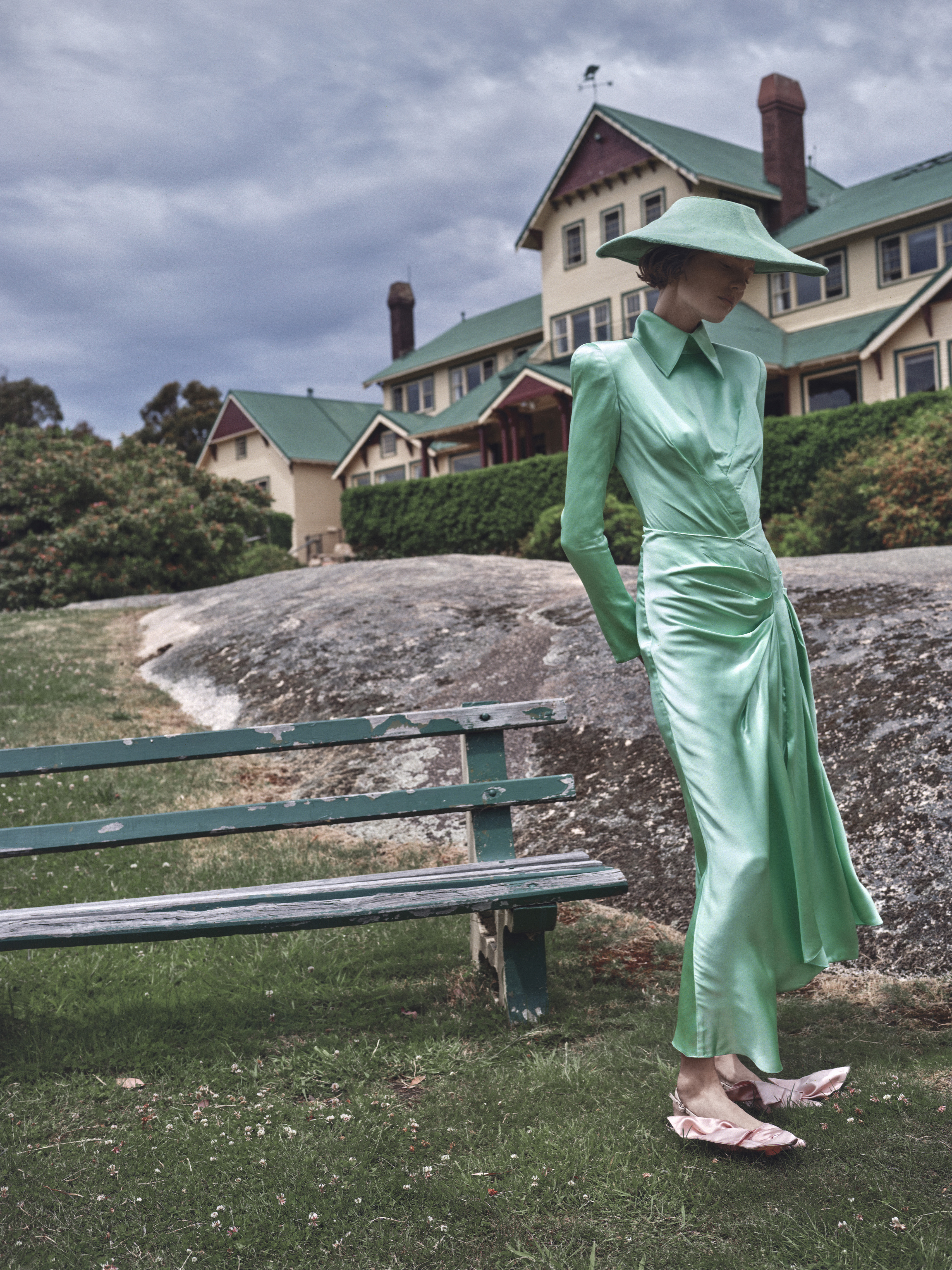 A woman in a green dress and hat stands on a grassy lawn near a bench, with a large building and cloudy sky in the background.