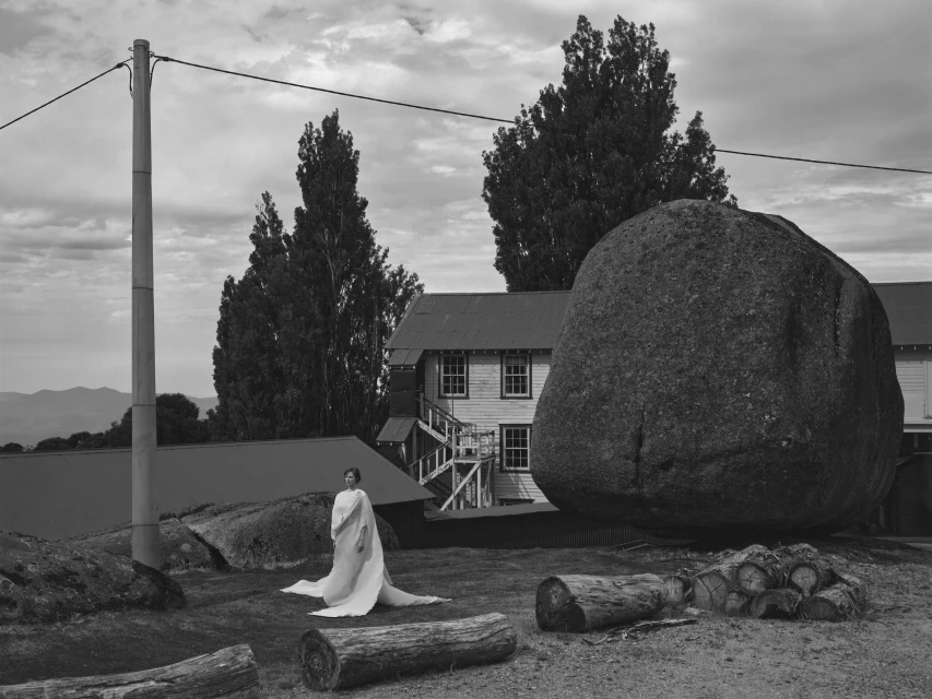 Monochromatic image of a woman in a white dress posing in a rural landscape with a large boulder, trees, and a building in the background.