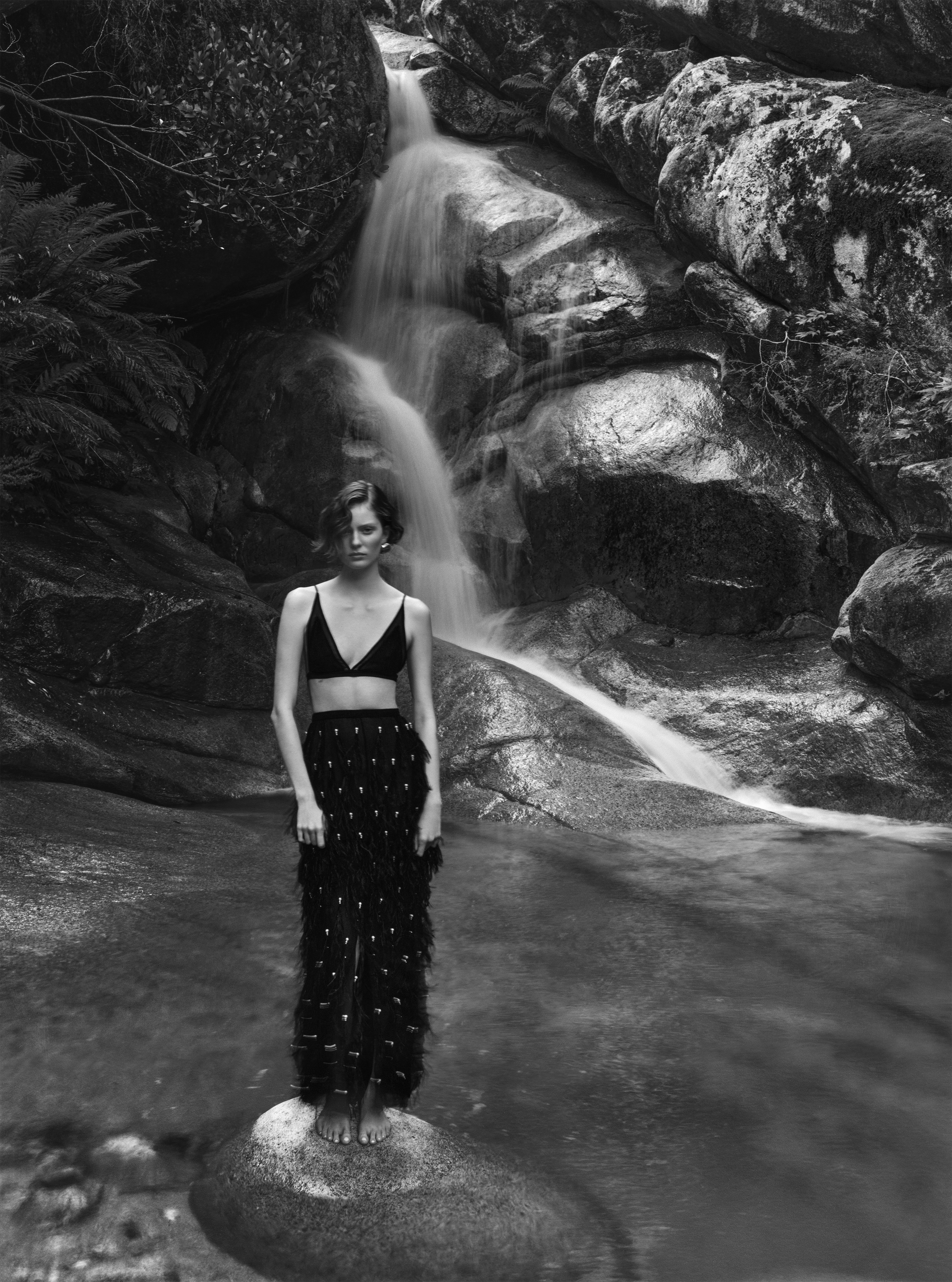 Woman in bra and skirt standing on rock in waterfall. Portrait of adult female posing in nature, black and white photography.