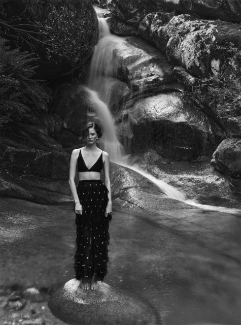 Woman in bra and skirt standing on rock in waterfall. Portrait of adult female posing in nature, black and white photography.