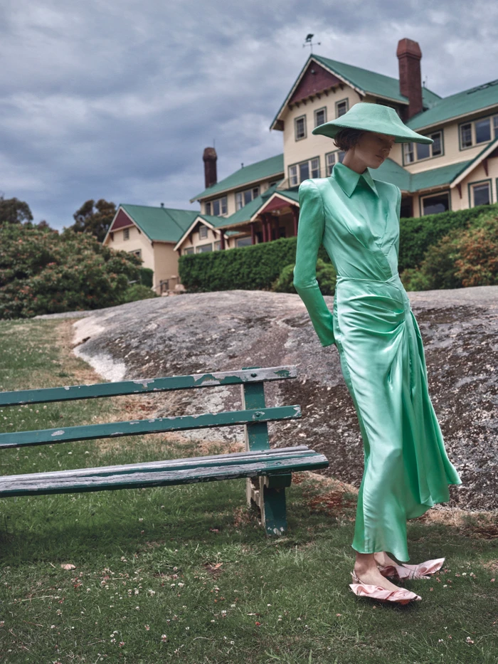 Model standing outdoors wearing a matching mint green satin dress and wide-brimmed hat in front of a large historic building.