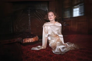 Woman in a glossy silk dress seated on a patterned red carpet inside a dimly lit rustic room