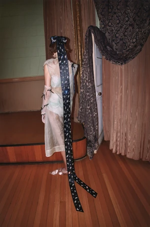 Model seen from behind wearing sheer dress and very long black hair ribbon in an interior room with draped curtains