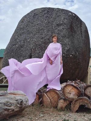 Model in a flowing light purple dress standing before a massive boulder and stacked logs for Vogue Polska.