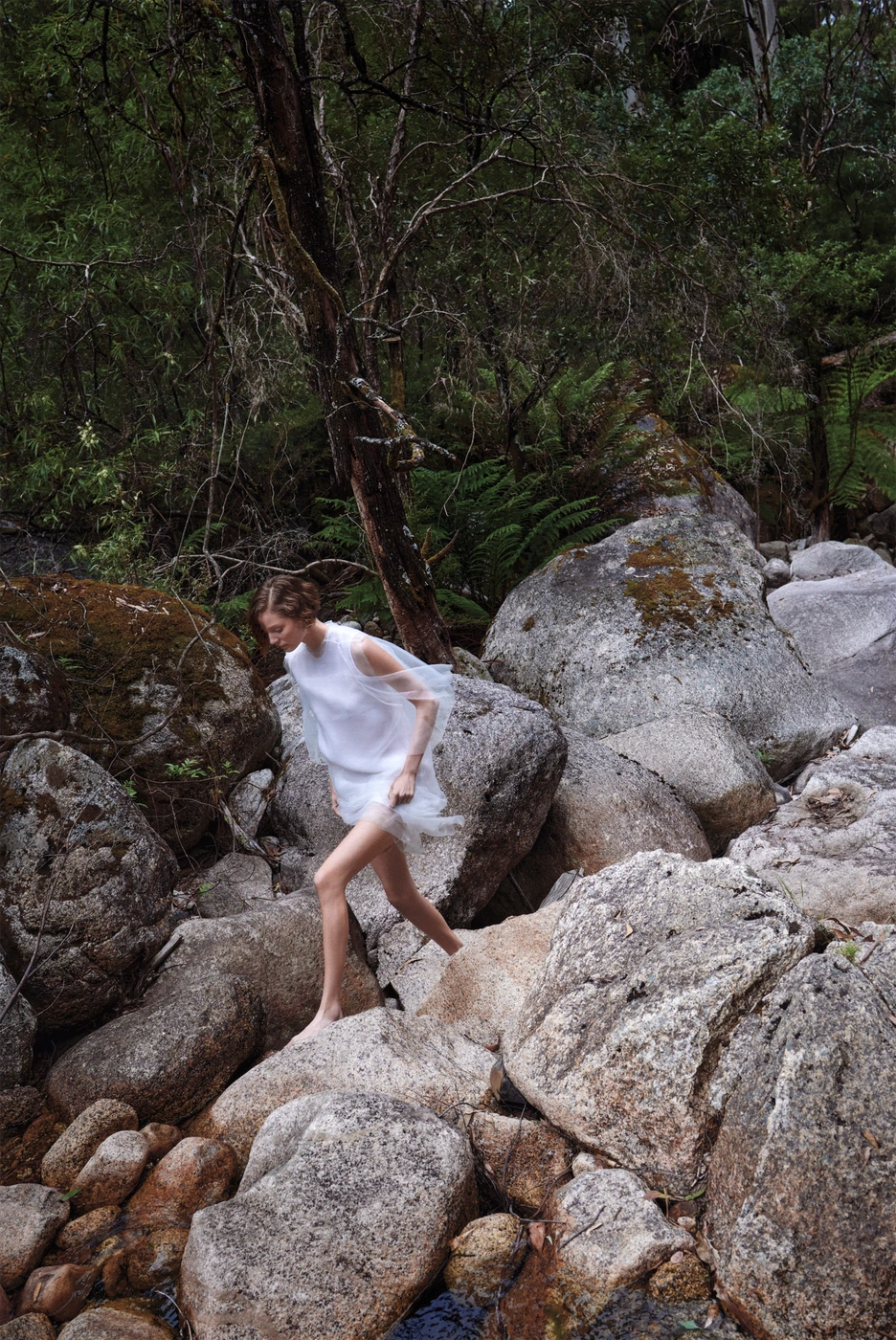 Model in sheer white dress stepping across large grey river boulders in a lush forest