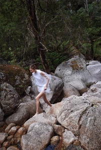 Model in sheer white dress stepping across large grey river boulders in a lush forest
