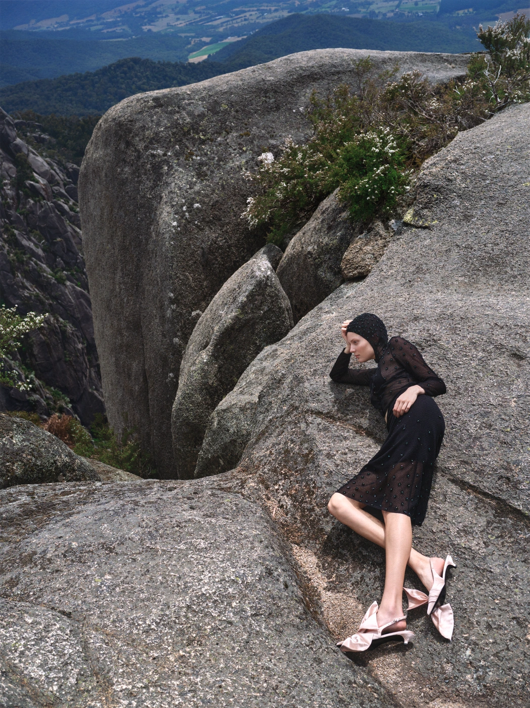 Model lying on a rugged mountain rock formation wearing a black sheer top, dark skirt, and pink heels with large bows.
