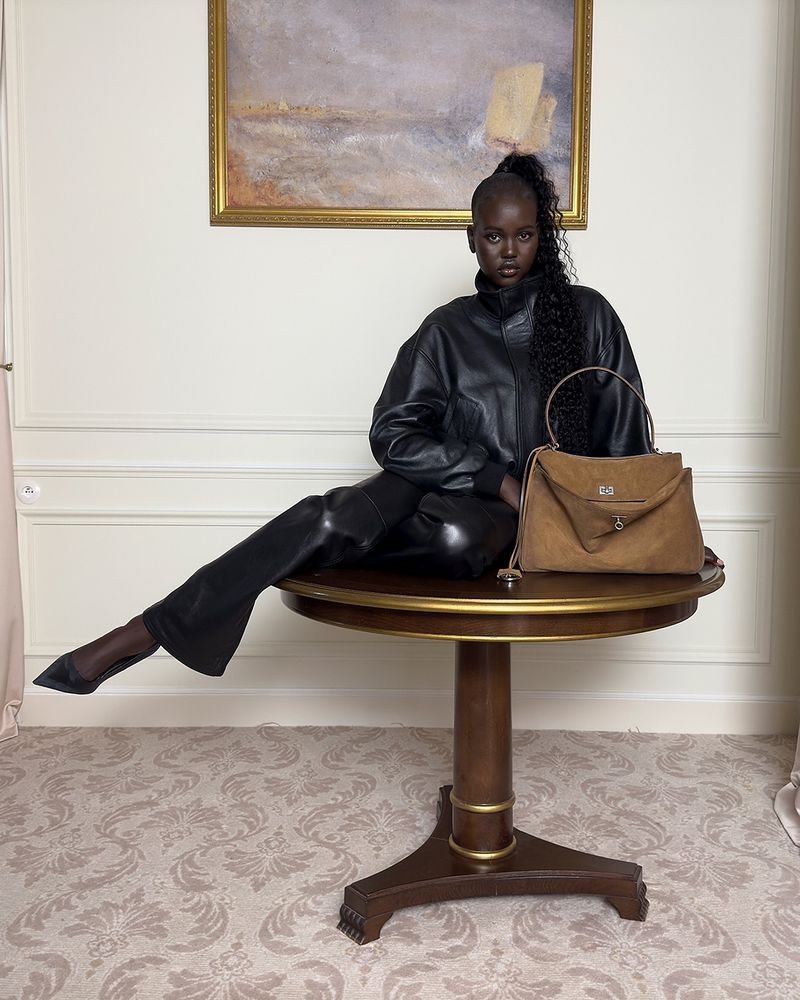 Elegant Black woman poses on a round wood table with a brown leather handbag in an indoor studio setting with cream colored walls.
