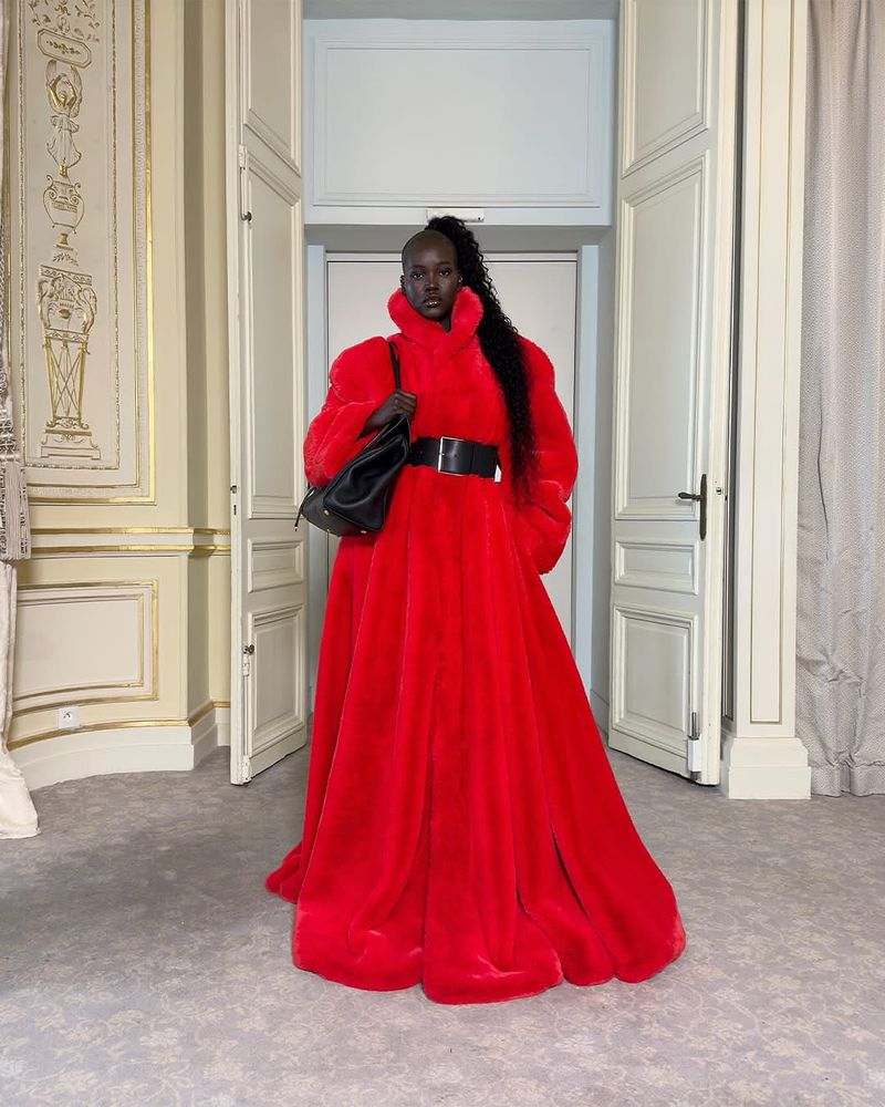 A dark-skinned female model in a long red coat with a black belt and handbag poses in a room with tall doors and moulding.