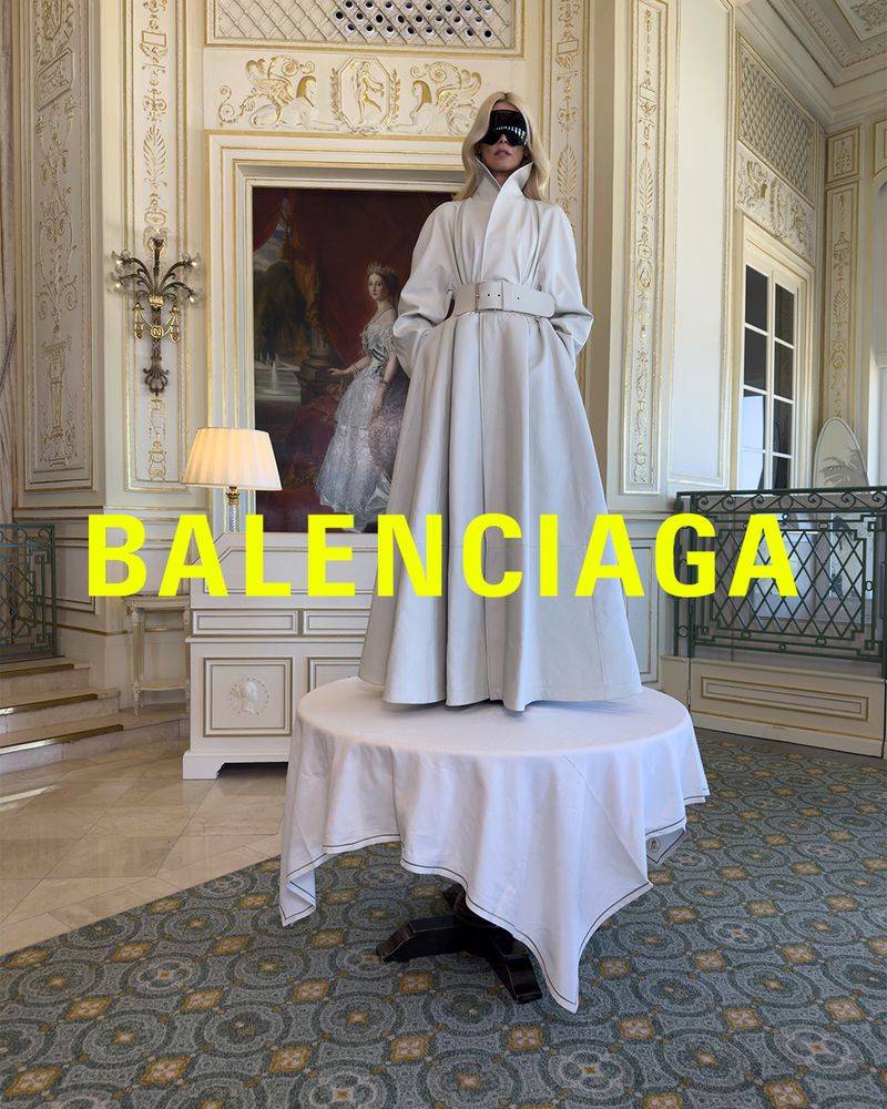 Blonde woman stands on table draped in white cloth wearing a white coat, black blindfold in room with decorative painting.