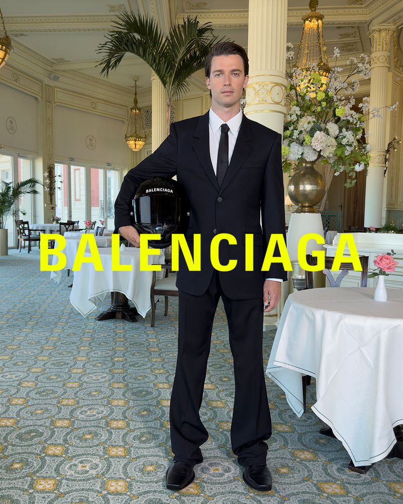 A man in a dark suit holds a helmet indoors, standing in front of a table setting with flowers and opulent architectural details.