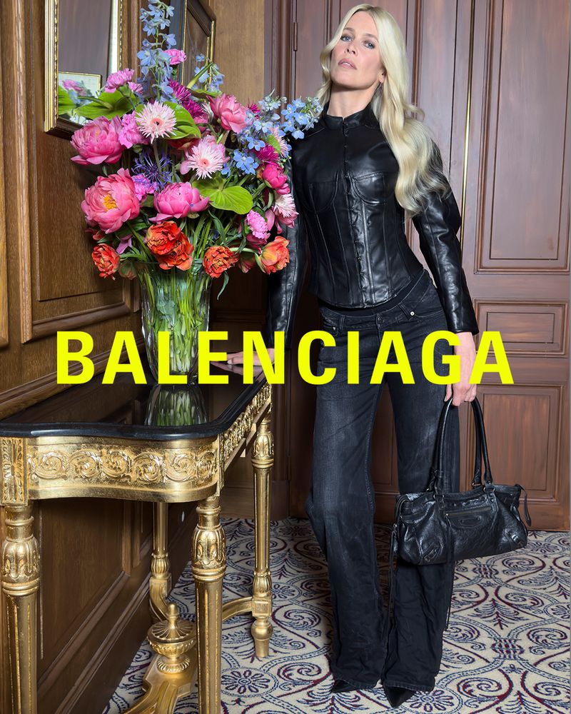 Model in black leather outfit and handbag poses next to flowers and gilded table for Balenciaga advertisement against paneled wall.